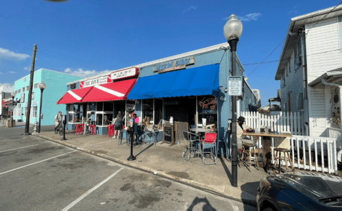 A colorful street scene featuring a restaurant with blue and red awnings, outdoor seating, and people enjoying the area.