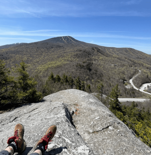 A hiker's feet on a rock ledge, overlooking a green mountain landscape under a clear blue sky.