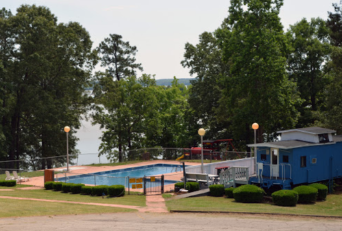 A serene outdoor pool area surrounded by trees, with a blue building and a view of a lake in the background.