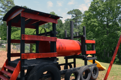 A colorful playground structure resembling a train, made of wood and tires, set in a grassy area with trees in the background.