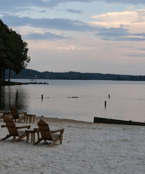 Adirondack chairs on a sandy beach by a calm lake at dusk, with trees and distant hills in the background.