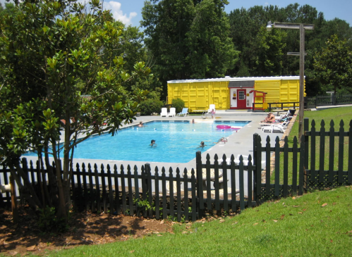 A sunny pool area with people swimming, surrounded by green grass and a yellow building in the background.