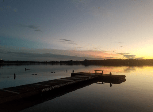 A serene lake at sunset, with a wooden dock extending into calm waters and colorful clouds in the sky.