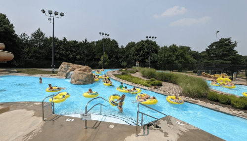 A lazy river at a water park with people floating on yellow tubes, surrounded by greenery and a clear blue sky.