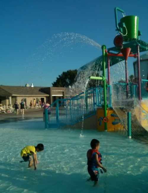 Two children play in a splash pad with water features and colorful structures under a clear blue sky.