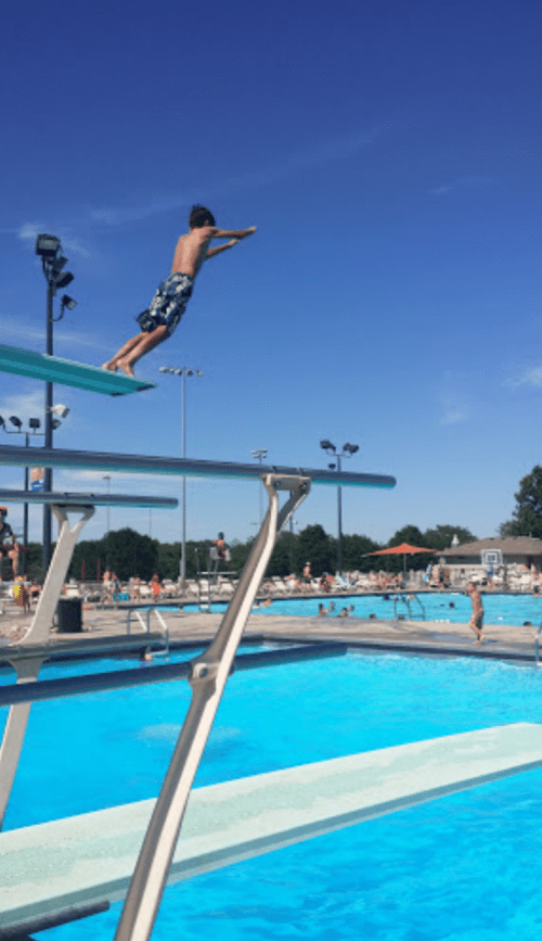 A boy prepares to dive from a high diving board into a bright blue swimming pool on a sunny day.