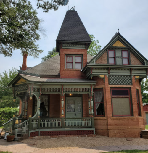 A charming Victorian-style house with a turret, intricate woodwork, and a wraparound porch surrounded by greenery.