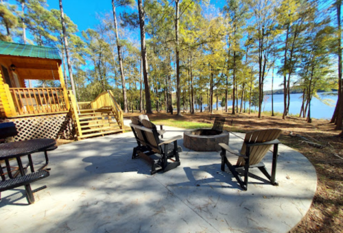 A serene outdoor space with a fire pit, wooden chairs, and a cabin surrounded by trees and a lake in the background.