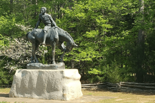 A bronze statue of a man on a horse, set against a backdrop of green trees and a wooden fence.
