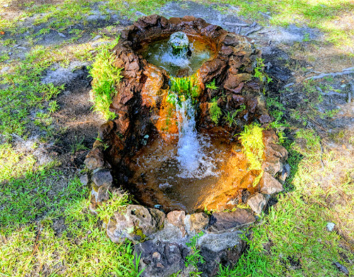 A small stone fountain surrounded by grass, with water flowing from the top into a basin below.