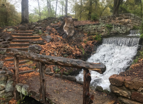 A wooden bridge over a small waterfall, surrounded by stone steps and autumn leaves in a serene forest setting.
