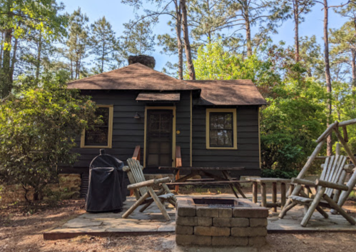 A cozy black cabin surrounded by trees, featuring a stone fire pit and wooden chairs in a serene outdoor setting.