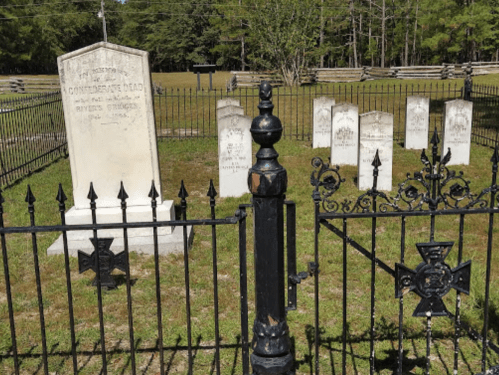 A historic cemetery with a prominent gravestone and several smaller markers, enclosed by a decorative black iron fence.