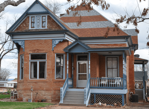 A historic brick house with blue trim, a porch, and a gabled roof, surrounded by trees and a cloudy sky.