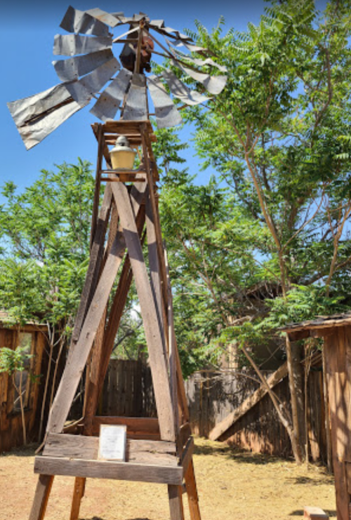 A rustic windmill stands tall among trees, with a wooden base and metal blades, in a sunny outdoor setting.