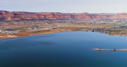 A serene lake reflects red rock formations and a small town, surrounded by fields and distant hills under a clear sky.