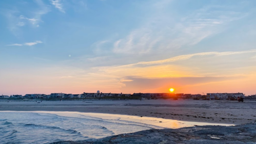 Sunset over a beach with gentle waves, reflecting warm colors in the sky and illuminating distant buildings along the shore.