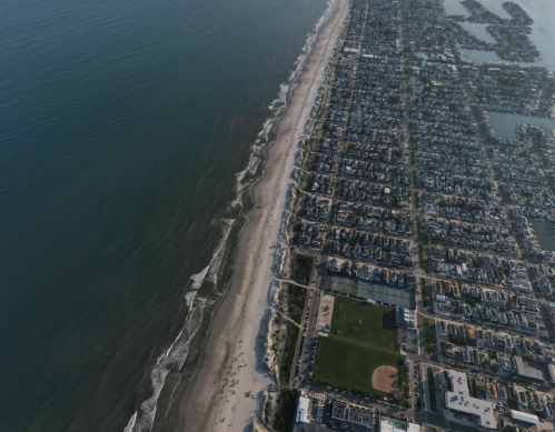 Aerial view of a coastline with a sandy beach, waves, and a nearby residential area with a baseball field.