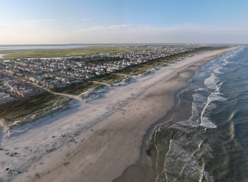 Aerial view of a sandy beach with gentle waves, bordered by a coastal town and lush green areas in the background.