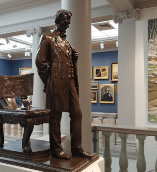 A bronze statue of Abraham Lincoln stands beside a wooden bench in an art gallery, surrounded by framed paintings.