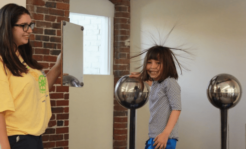 A child with wild hair smiles while standing next to a woman holding a mirror in a science exhibit.