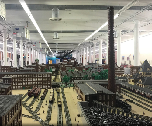 A detailed model of a factory town with buildings, trees, and train tracks, set in a spacious indoor environment.