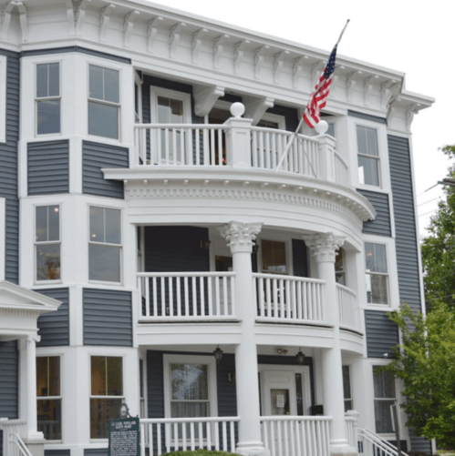 A historic building with a curved balcony, white columns, and an American flag, featuring blue and white siding.