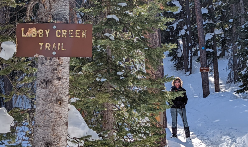A person stands on a snowy trail next to a sign for Lobby Creek Trail, surrounded by trees.