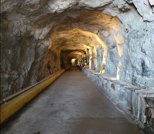 A dimly lit tunnel with rocky walls and a smooth concrete floor, leading to a bright opening in the distance.