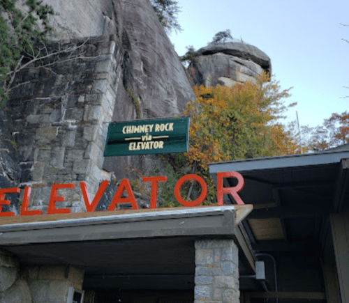 Sign for "Chimney Rock via Elevator" with a rocky cliff and autumn foliage in the background.