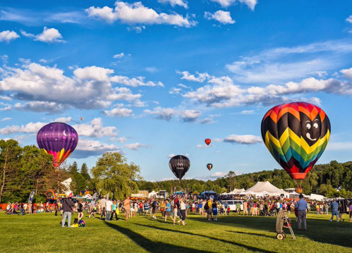 Colorful hot air balloons float above a lively crowd at a festival on a sunny day with blue skies and fluffy clouds.