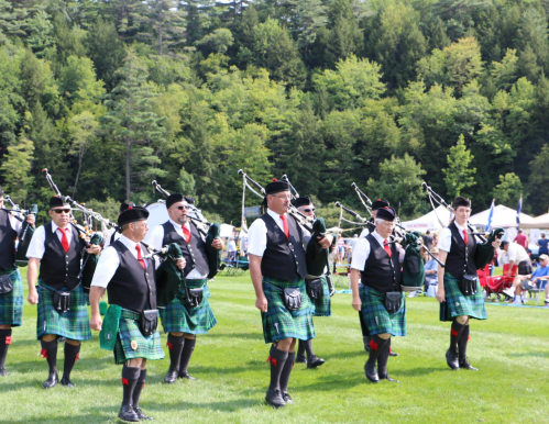 A group of bagpipers in traditional attire march on a grassy field, surrounded by trees and spectators.