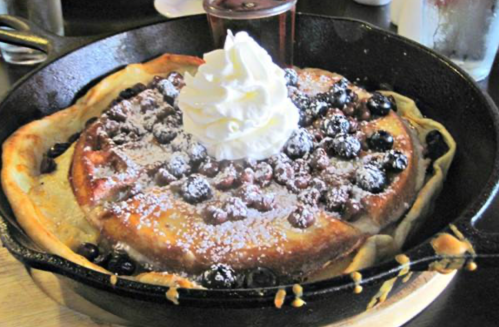 A skillet filled with a fluffy pancake topped with whipped cream, blueberries, and powdered sugar.