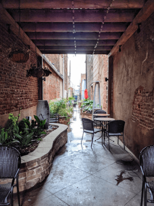 A narrow, brick-lined alleyway with potted plants, a table and chairs, and a wet pavement reflecting light.