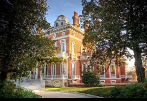 A grand brick mansion with ornate architecture, surrounded by lush greenery and trees, under a clear blue sky.