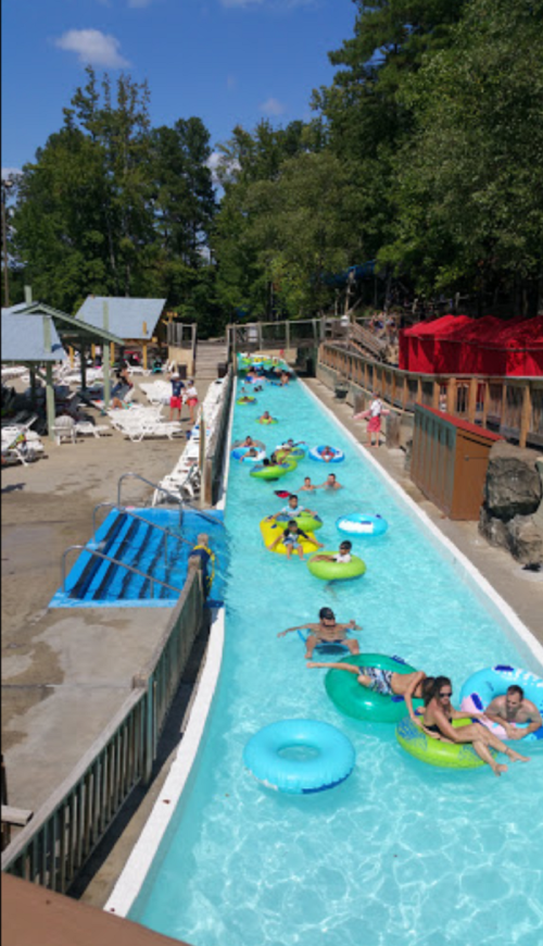 A lazy river at a water park with colorful inner tubes and people enjoying the sun. Trees and lounge chairs in the background.