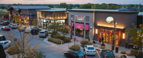 A shopping plaza at dusk with various stores, parked cars, and trees lining the walkways.