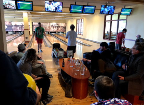 A bowling alley scene with people sitting on a couch, watching a bowler, and several screens displaying scores.