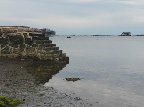 Stone steps lead down to calm water, with a distant island visible under a cloudy sky.