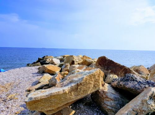 Rocky shoreline with large boulders, leading to a calm sea under a clear blue sky.