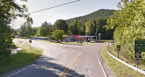 A rural intersection with a gas station, surrounded by trees and mountains, under a clear blue sky.