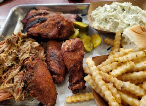 A metal tray with fried chicken, pulled pork, pickles, crinkle-cut fries, coleslaw, and a bun.