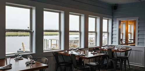 Cozy restaurant interior with wooden tables, chairs, and large windows overlooking a scenic waterfront view.