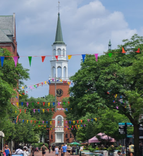 A vibrant street scene with colorful flags, trees, and a clock tower in the background under a blue sky.