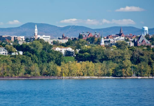 A scenic view of a city skyline with mountains in the background, featuring greenery and a calm body of water in the foreground.