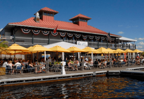 A waterfront restaurant with outdoor seating under yellow umbrellas, bustling with diners on a sunny day.