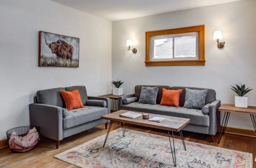 Cozy living room with gray sofas, orange cushions, a wooden table, and a wall art of a bison. Bright and inviting atmosphere.