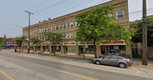 A street view of a brick building with storefronts and trees lining the sidewalk, parked cars visible.