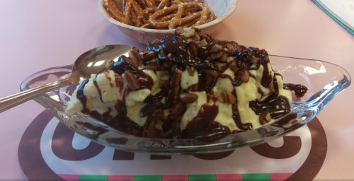 A glass dish of ice cream topped with chocolate sauce and nuts, alongside a bowl of pretzels.
