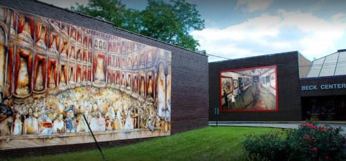 Two large murals on a brick building, depicting vibrant scenes, next to the entrance of the Beck Center. Green grass in front.